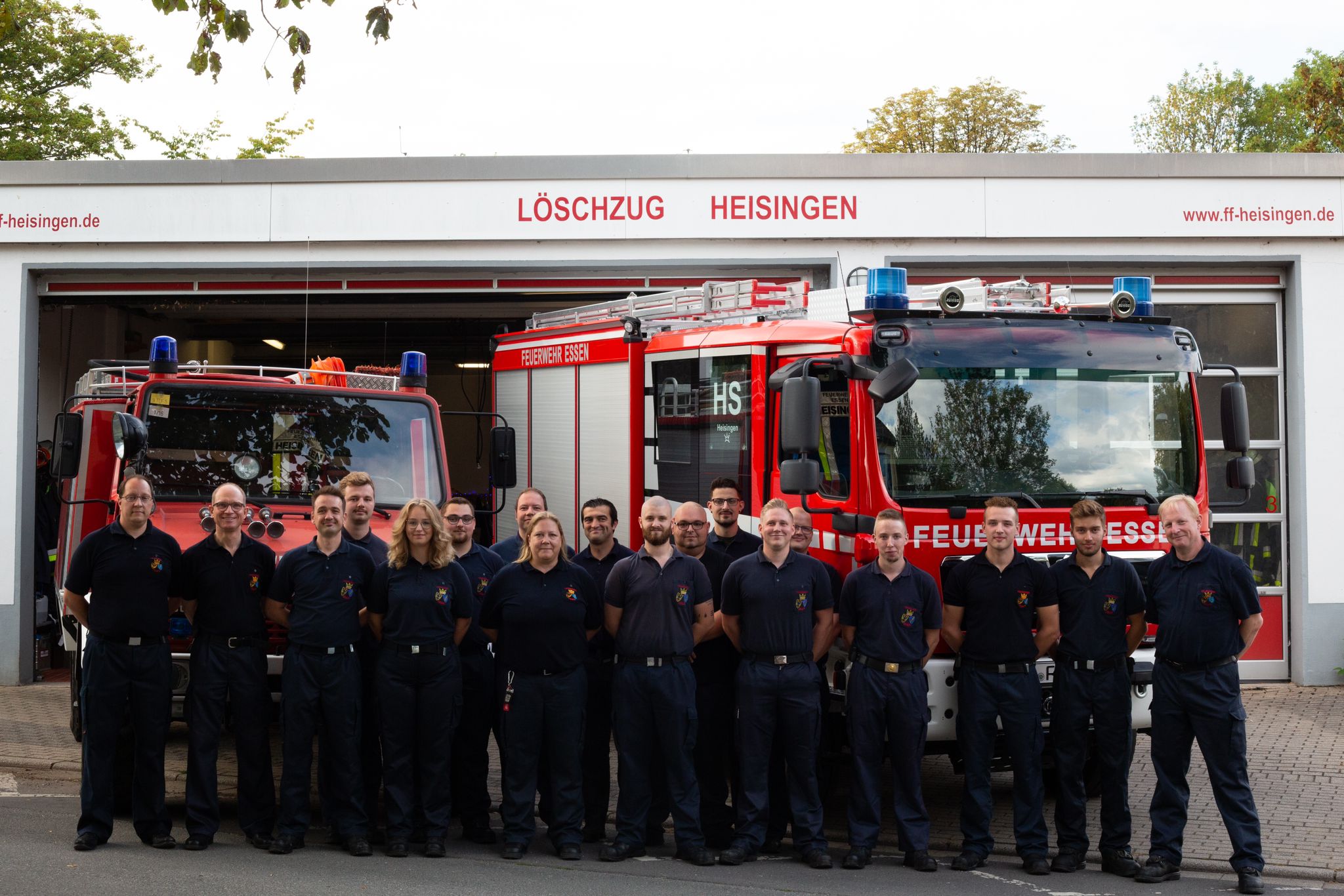 Mannschaftsfoto der Freiwilligen Feuerwehr Essen Heisingen vor dem Gerätehaus in der Zölestinstraße. Die Mannschaft steht vor den Einsatzfahrzeugen: Einem Tanklöschfahrzeug und einem Hilfeleistungslöschfahrzeug.